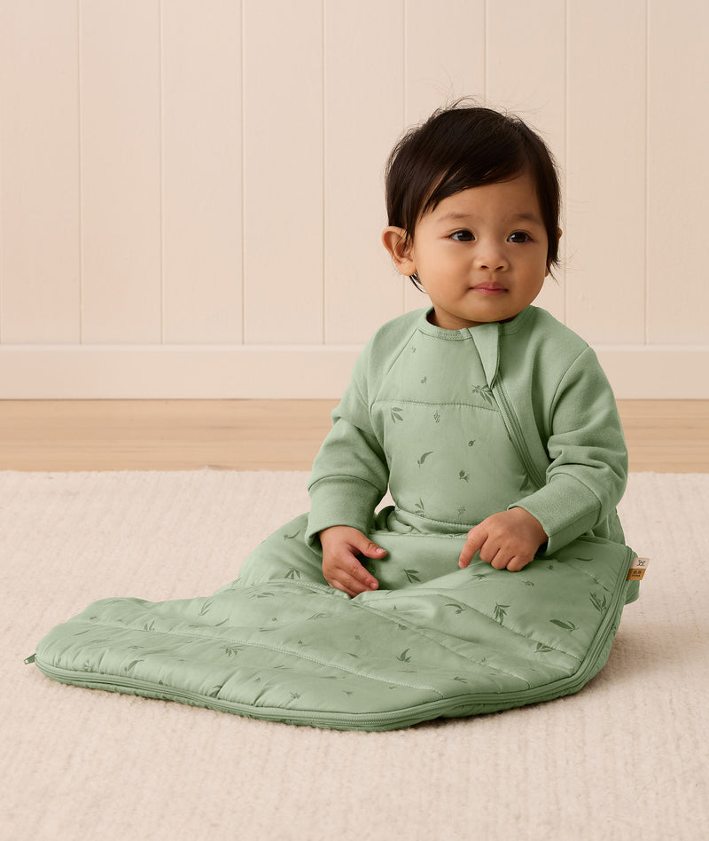 An infant sitting on a rug is wearing an ergoPouch Premium Sleeping Bag featuring side zippers.
