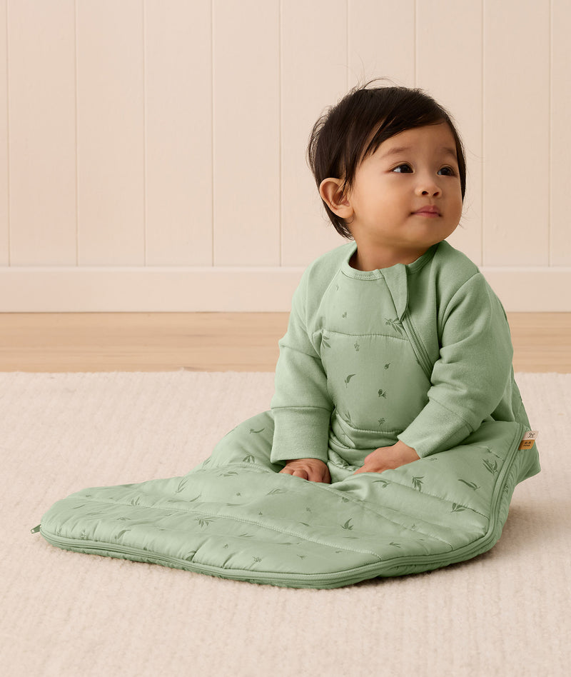 An infant sitting on a rug is wearing an ergoPouch Premium Sleeping Bag featuring side zippers.