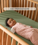 Baby lying in a cot with a green mattress and wooden sides, wearing a pink sleeping bag.