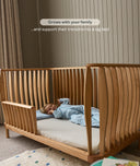 Wooden cot with a child lying inside, set against a neutral wall.