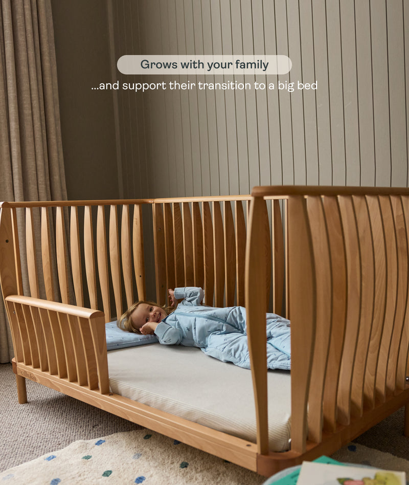 Wooden cot with a child lying inside, set against a neutral wall.