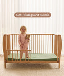 Child standing in a wooden cot with a green mattress and sideguard, with text 'Cot + Sideguard bundle' above.