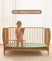 Child standing in a wooden cot with a green mattress and sideguard, with text 'Cot + Sideguard bundle' above.