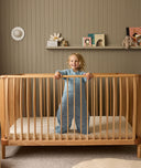 Child standing in a wooden cot with a shelf of toys and decorations in the background.