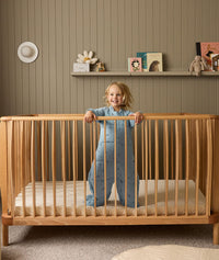 Child standing in a wooden cot with a shelf of toys and decorations in the background.