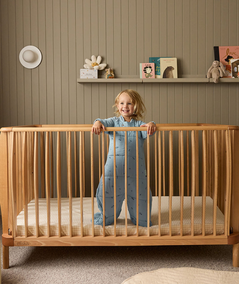 Child standing in a wooden cot with a shelf of toys and decorations in the background.