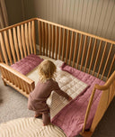 Child standing next to a wooden cot with pink bedding in a room with wooden paneling.