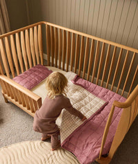 Child standing next to a wooden cot with pink bedding in a room with wooden paneling.