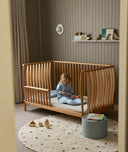 Child sitting in a wooden cot in a room with a shelf and toys on the floor.