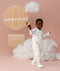Preschooler wearing a white Sleep Onesie with a colourful print, walking against a soft pink background and smiling while holding a toy plane, with cloud props and a hot air balloon illustration behind them.