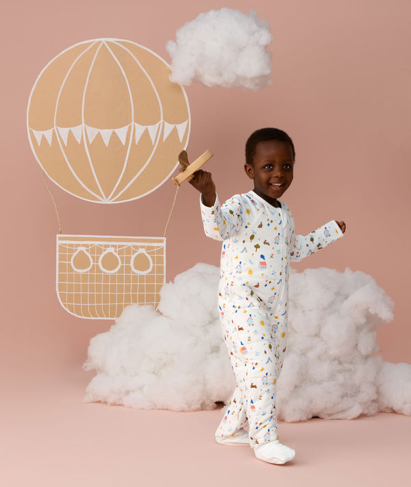 Preschooler wearing a white Sleep Onesie with a colourful print, walking against a soft pink background and smiling while holding a toy plane, with cloud props and a hot air balloon illustration behind them.