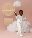Preschooler wearing a white Sleep Onesie with a colourful print, walking against a soft pink background and smiling while holding a toy plane, with cloud props and a hot air balloon illustration behind them.