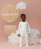 Preschooler wearing a white sleep onesie with a colourful print, standing against a soft pink background and smiling, with cloud props and a hot air balloon illustration behind them.