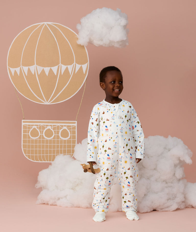 Preschooler wearing a white sleep onesie with a colourful print, standing against a soft pink background and smiling, with cloud props and a hot air balloon illustration behind them.