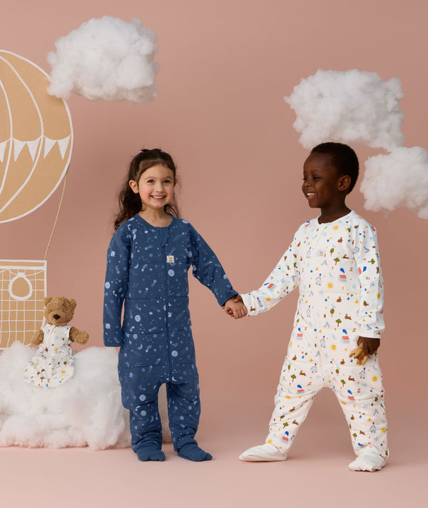 Two preschoolers holding hands against a soft pink background, one wearing a blue sleep onesie with a moon-and-star print and the other wearing a white sleep onesie with a colourful print, surrounded by cloud props and a hot air balloon illustration.