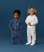 Two toddlers standing on a blue background wearing ergoPouch Sleep Onesie from the Dreamers collection. One wearing a blue onesie with star and moon print, while the other wearing a white onesie with colourful print.