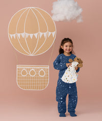 Preschooler wearing a blue Sleep Onesie with a moon-and-star print, standing against a soft pink background and smiling while holding a teddy bear, with a hot air balloon illustration behind them.
