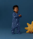 Toddler wearing a blue Sleep Onesie 2.5 TOG with star and moon all-over print, standing against a blue background with a sun plush pillow on the floor