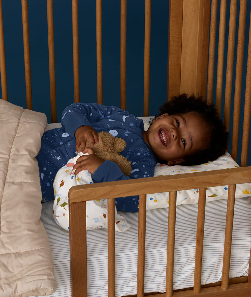Toddler lying in a wooden cot wearing a blue Sleep Onesie with a moon-and-star print, smiling and cuddling a teddy bear under a quilted blanket.