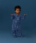Toddler wearing a blue Sleep Onesie with a moon-and-star print, standing against a deep blue background with arms raised and looking slightly to the side.