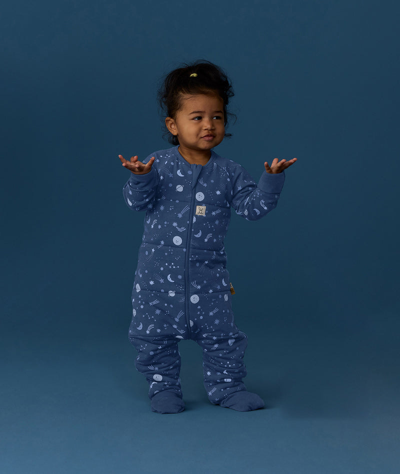 Toddler wearing a blue Sleep Onesie with a moon-and-star print, standing against a deep blue background with arms raised and looking slightly to the side.