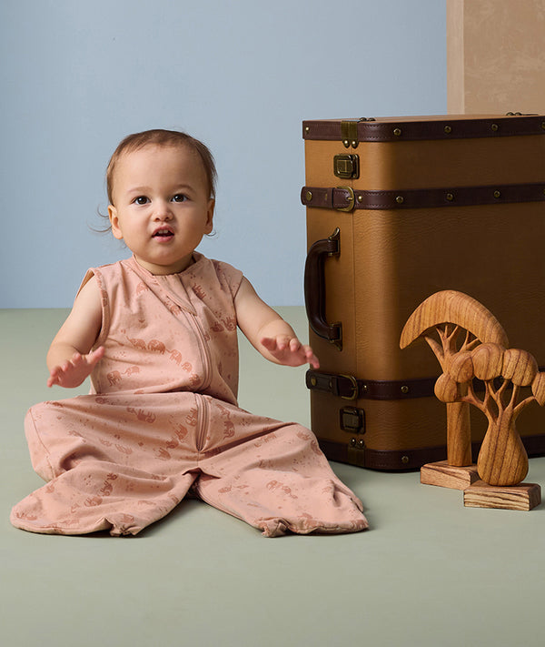 A toddler sits on a soft green floor, wearing a pink sleeveless sleep suit bag with a subtle animal print featuring elephants. The sleep suit has a front zipper and foot openings for mobility.