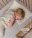 Child sleeping in a wooden cot on a matching patterned pillow, wearing a sleeveless cream Sleep Suit Bag.