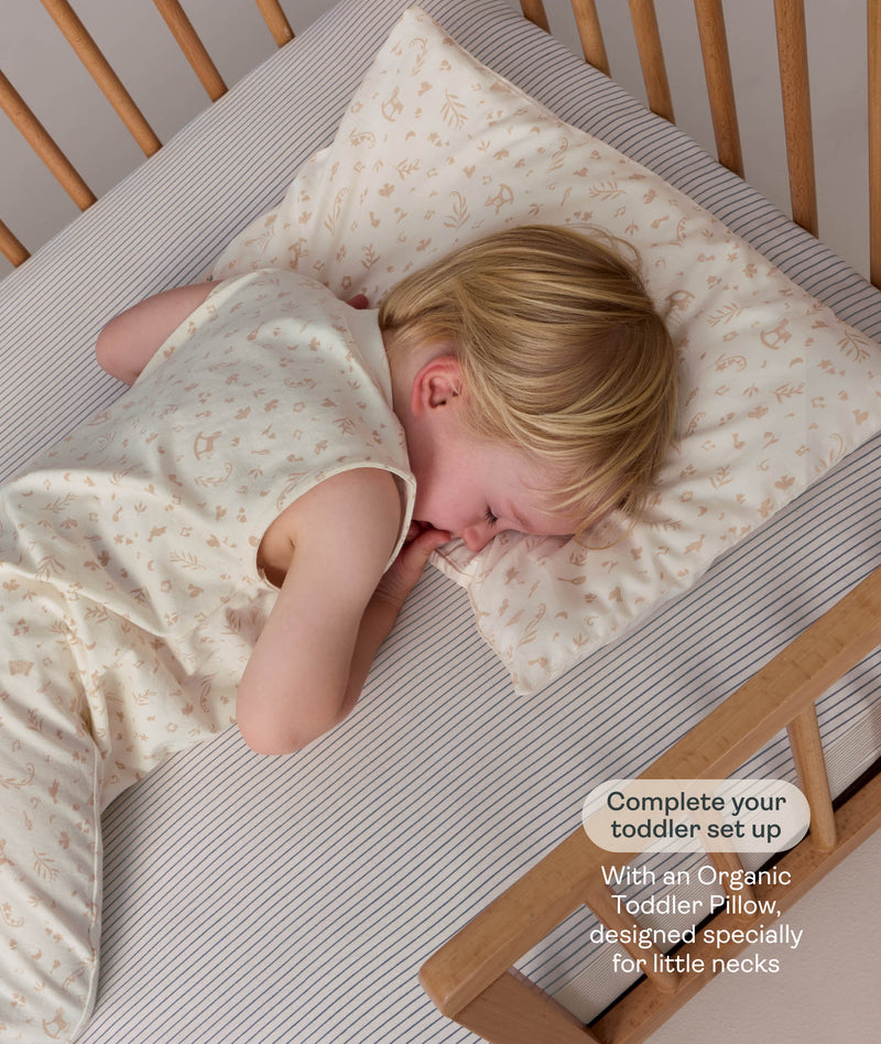 Child sleeping in a wooden cot on a matching patterned pillow, wearing a sleeveless cream Sleep Suit Bag.