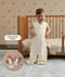 A smiling toddler standing next to a crib wearing a sleeveless, cream-patterned sleep suit bag in suit mode with legs with text highlighting matching doll sleeping bag.
