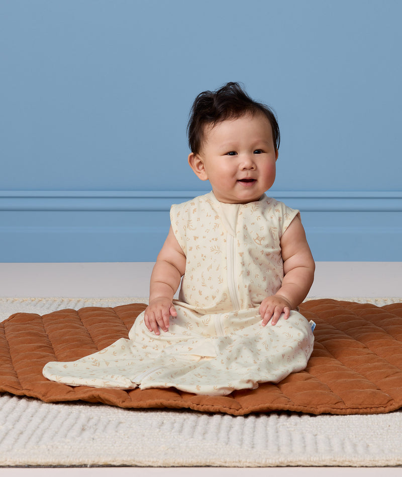 Infant sitting on a brown mat, wearing a cream sleeveless Sleep Suit Bag 0.2 TOG.