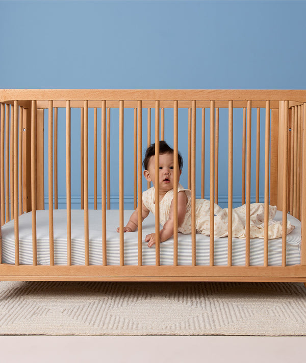 Baby lying on their tummy in a wooden cot wearing a sleeveless cream printed Sleep Suit Bag.