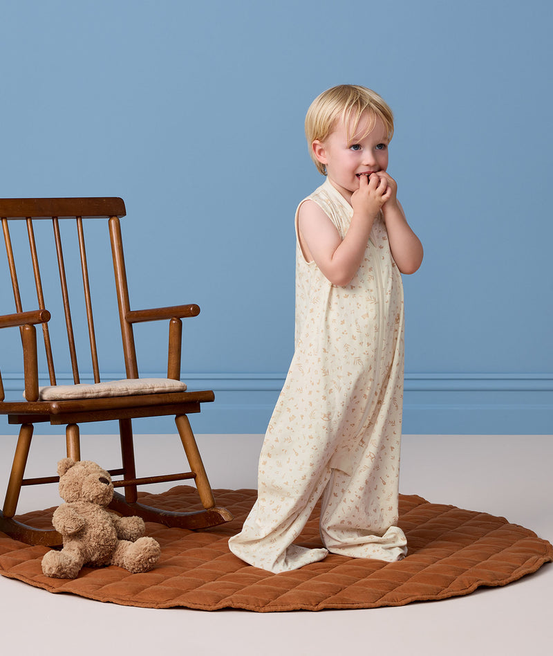 Toddler standing next to a wooden rocking chair wearing an ergoPouch  Sleep Suit Bag 0.2 TOG made from organic cotton.