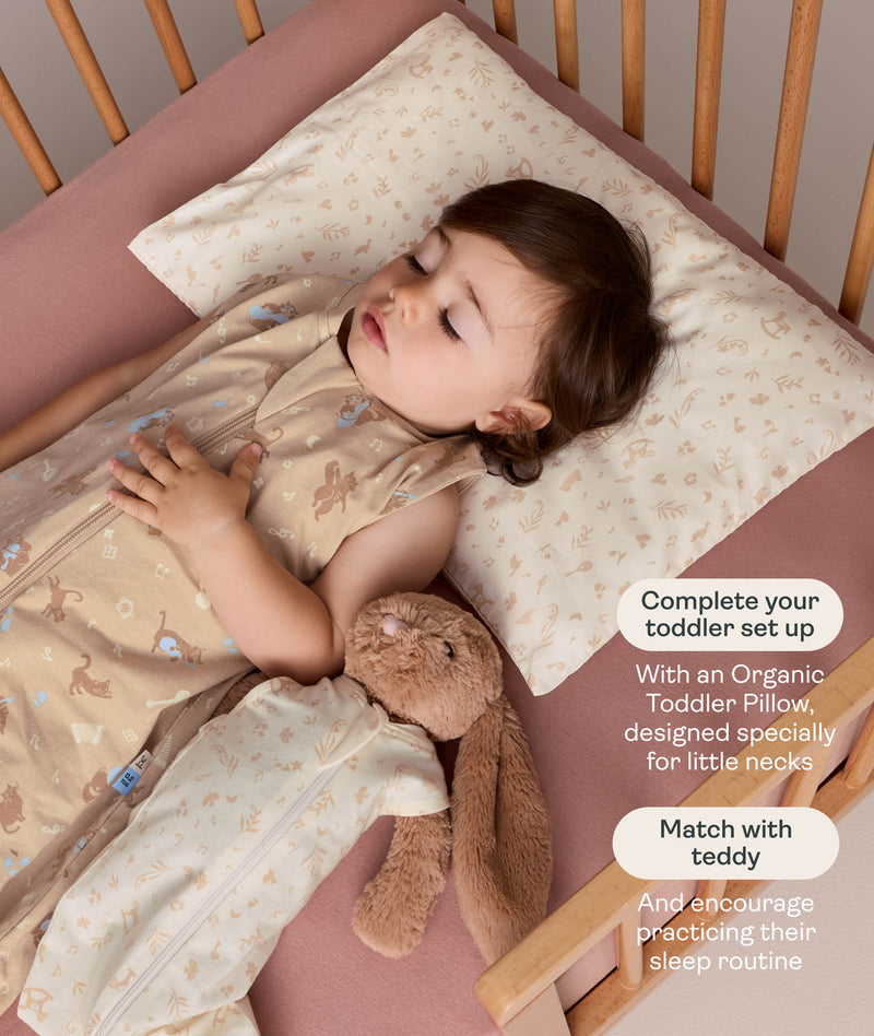 A sleeping toddler in a wooden cot, resting their head on a patterned toddler pillow while wearing a tan sleep suit next to a plush bunny dressed in a matching mini sleeping bag.