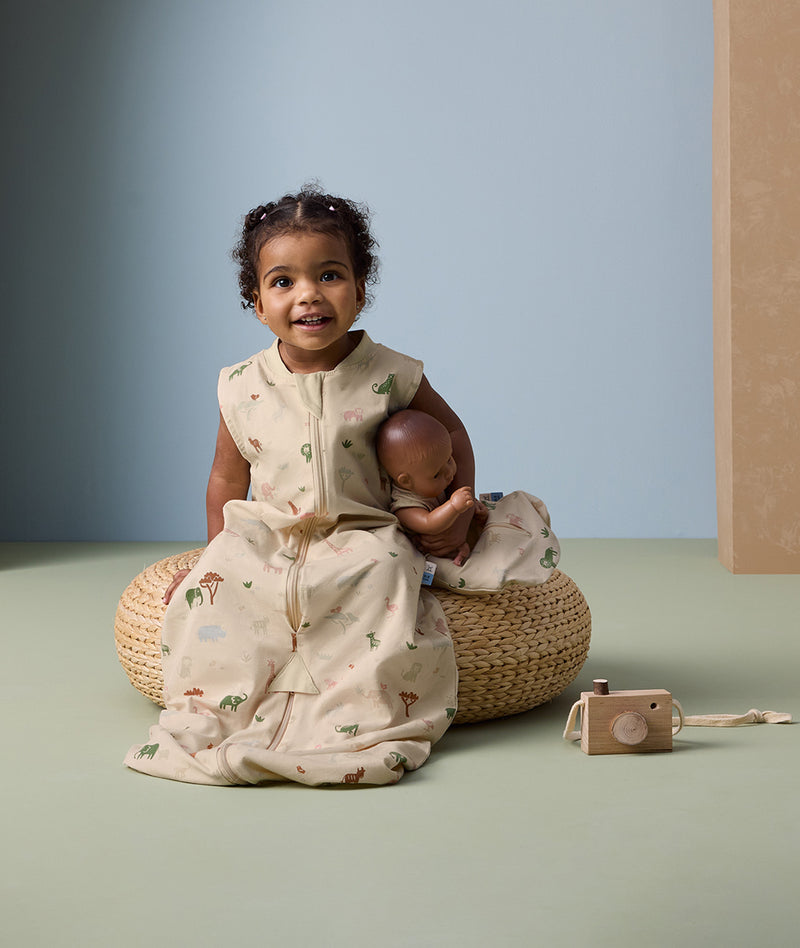 Toddler sitting on a woven cushion, wearing a safari-themed sleep suit bag and holding a baby doll dressed in a matching doll-sized sleeping bag.