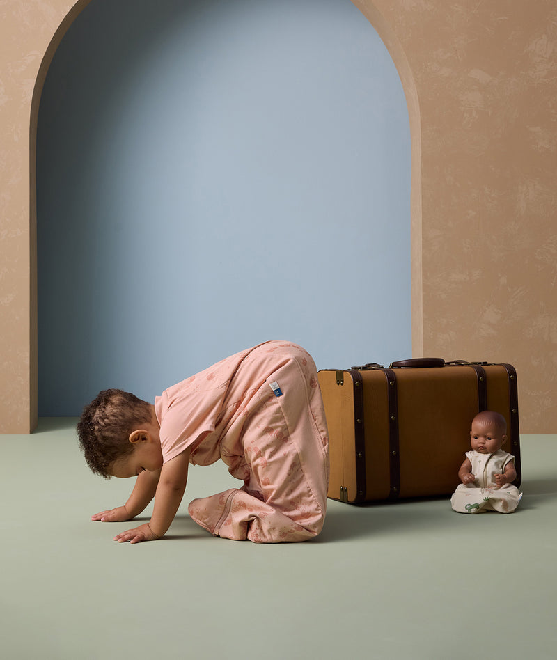 A toddler wearing a peach-colored sleep suit bag with a subtle animal print is seen in a playful crawling pose on a soft green floor. The sleep suit features short sleeves and a cozy, roomy fit for movement.