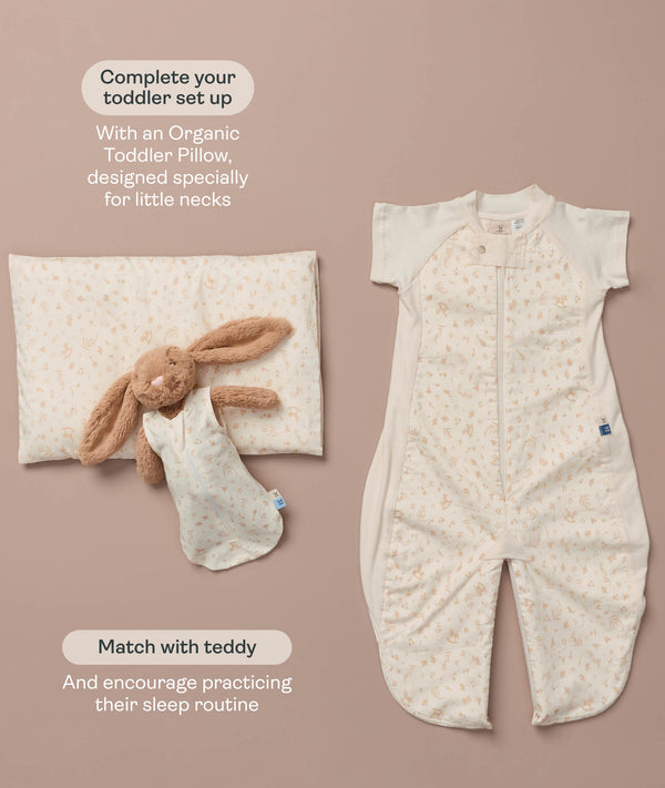 A cream printed toddler sleep suit with legs laid flat alongside a matching toddler pillow and a plush bunny wearing a mini sleeping bag.