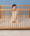 Baby standing in a wooden crib wearing a cream patterned Sleep Suit Bag with legs.