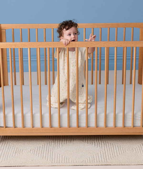 Baby standing in a wooden crib wearing a cream patterned Sleep Suit Bag with legs.