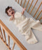 Baby reaching up while lying in a wooden cot, wearing a convertible sleep suit bag with leg openings.