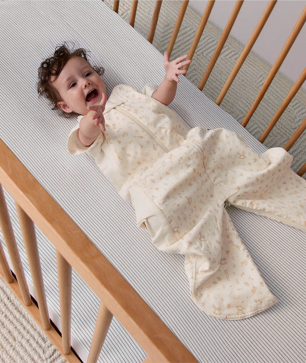 Baby reaching up while lying in a wooden cot, wearing a convertible sleep suit bag with leg openings.