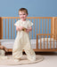 Toddler standing in front of a toddler bed, wearing a cream Sleep Suit Bag in suit mode while holding a wooden toy.