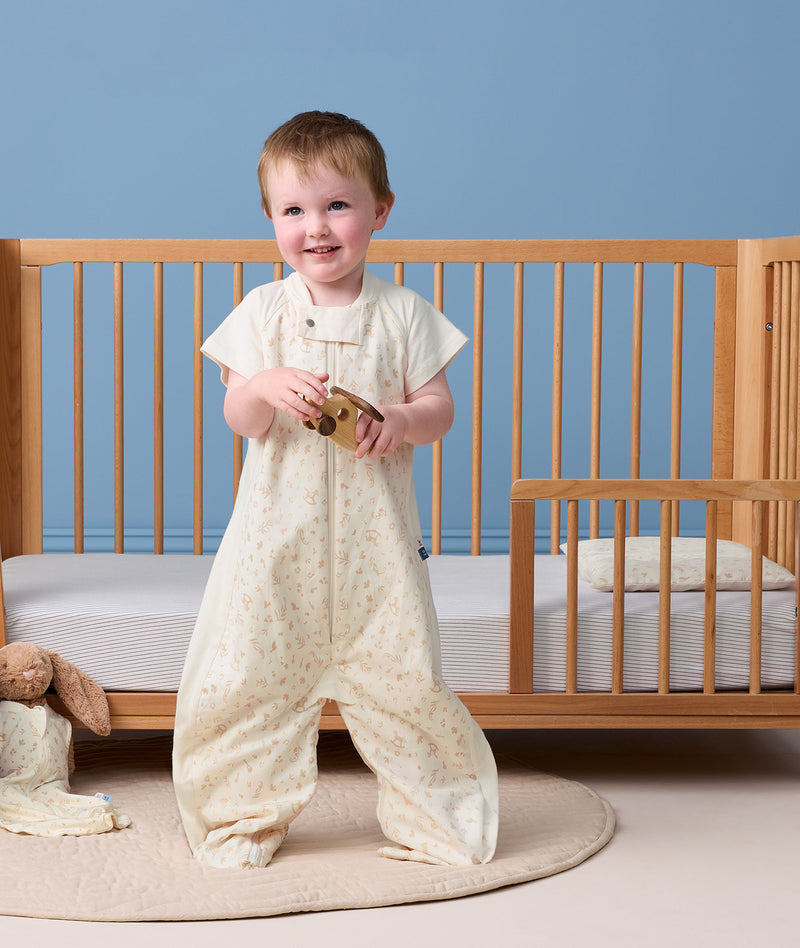 Toddler standing in front of a toddler bed, wearing a cream Sleep Suit Bag in suit mode while holding a wooden toy.