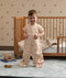 A smiling toddler standing on a rug in front of a wooden toddler bed, wearing a tan patterned convertible sleep suit in suit mode with legs.