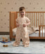 A smiling toddler standing on a rug in front of a wooden toddler bed, wearing a tan patterned convertible sleep suit in suit mode with legs.