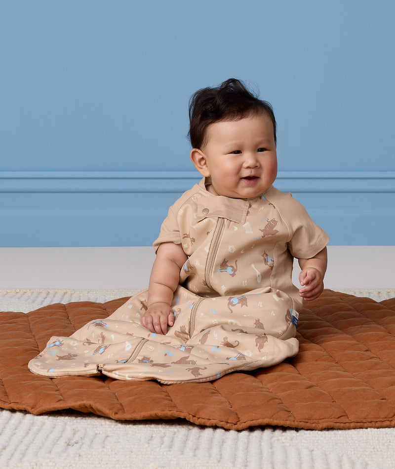 An infant sitting on a brown mat, wearing an ergoPouch Sleep Suit Bag 1.0 TOG made from premium cotton fabric.