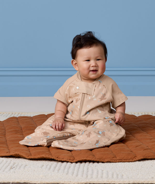 An infant wearing a short-sleeved Sleep Suit Bag 1.0 TOG in suit mode, is sitting on a brown quilted mat.