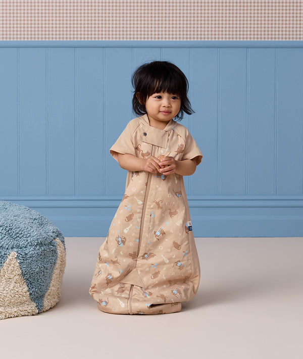 A toddler standing while wearing a tan short-sleeved Sleep Suit Bag in sleeping bag mode.