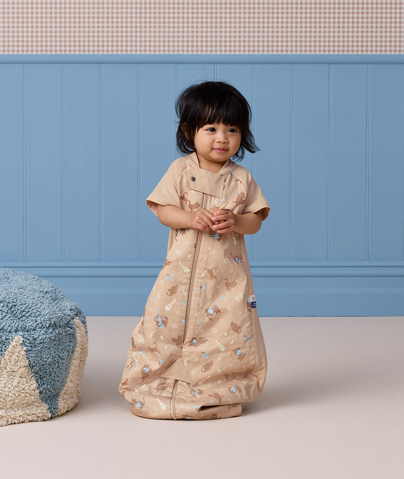A toddler standing while wearing a tan short-sleeved Sleep Suit Bag in sleeping bag mode.