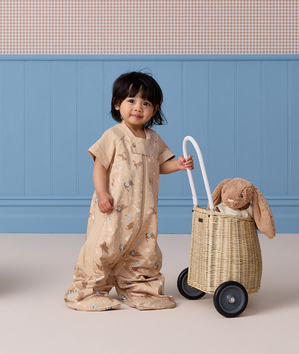 A toddler standing next to a toy cart wearing a tan short-sleeved convertible sleep suit bag in suit mode.