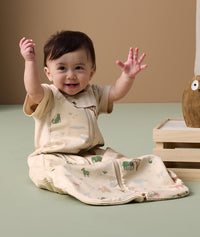The infant sits on the floor, raising both hands while wearing an ergoPouch Sleep Suit Bag in Savannah.